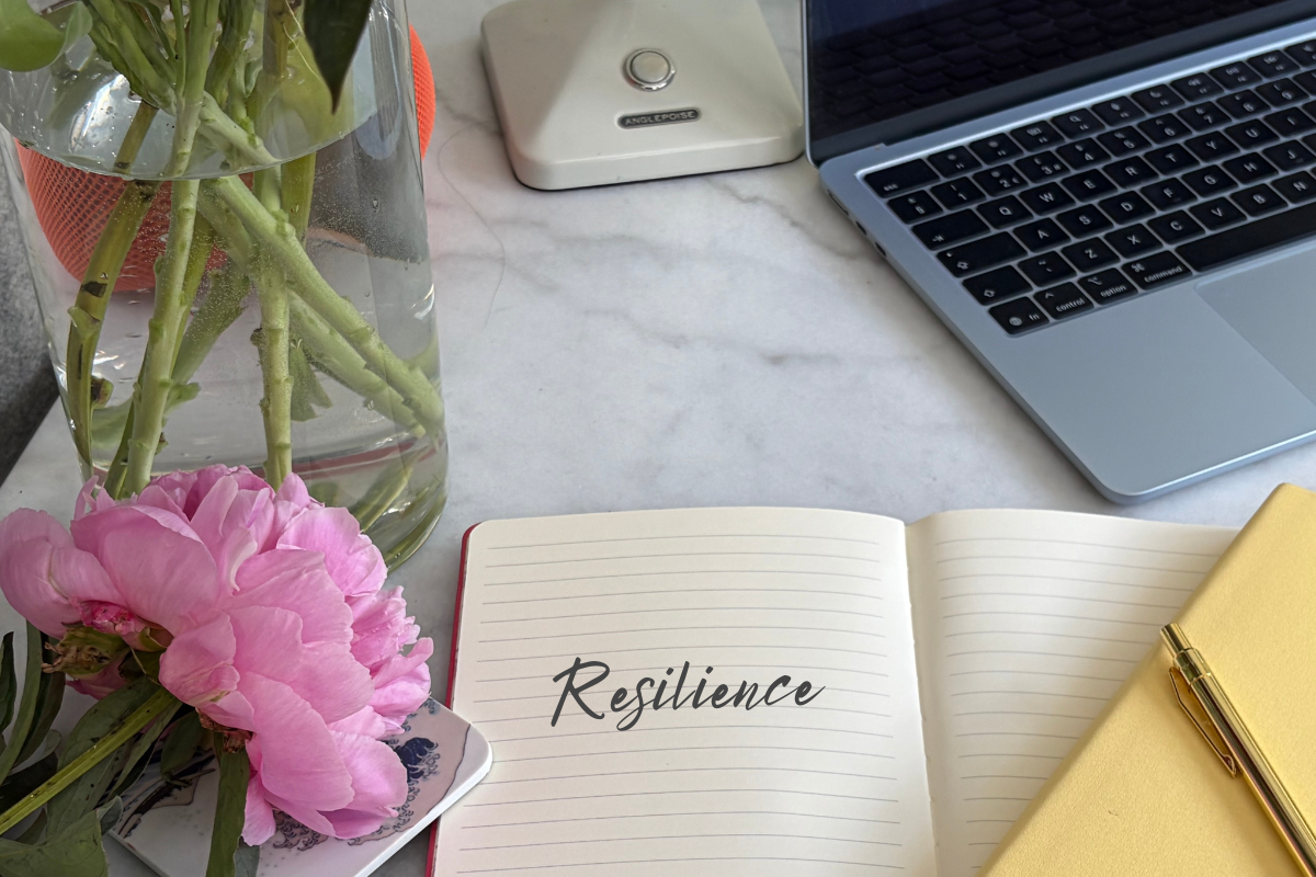 Open notebook with 'Resilience' handwritten in cursive, next to a pink peony in a glass vase, a laptop and a pen on a marble desk.